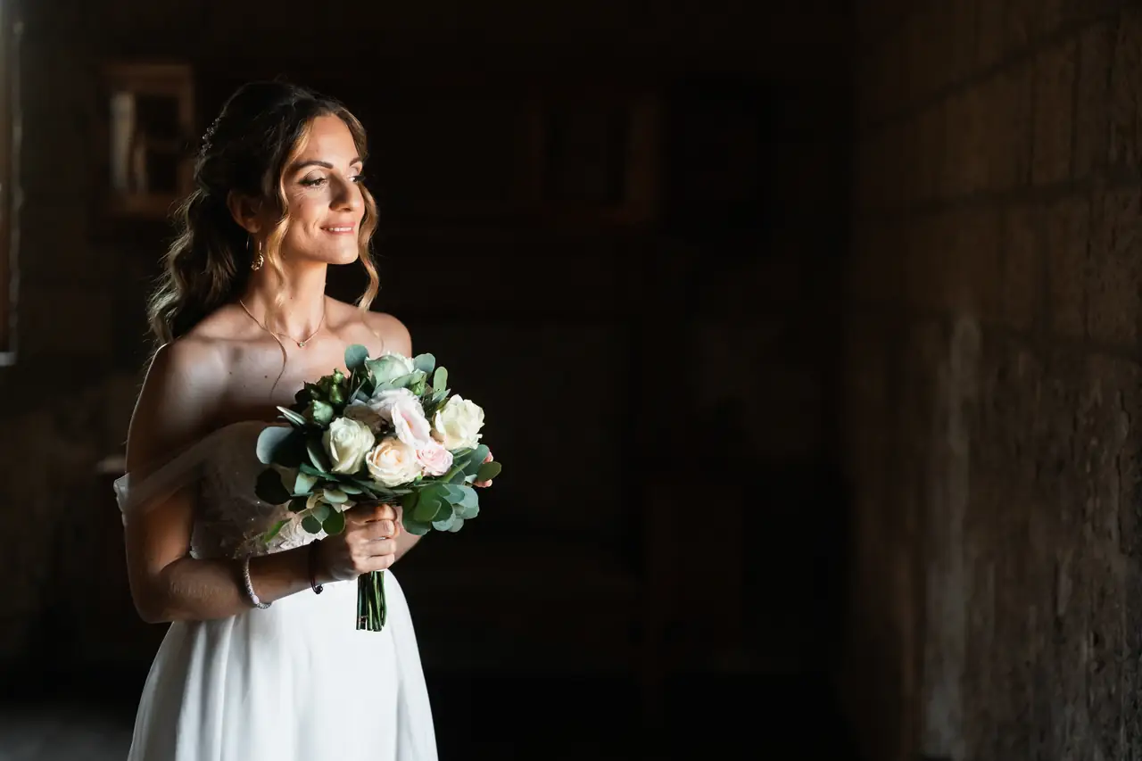 La sposa, in un elegante abito bianco con le spalle scoperte, tiene in mano uno splendido bouquet del rinomato fiorista Il Petalo di Marino, che risplende con grazia in una luce soffusa.