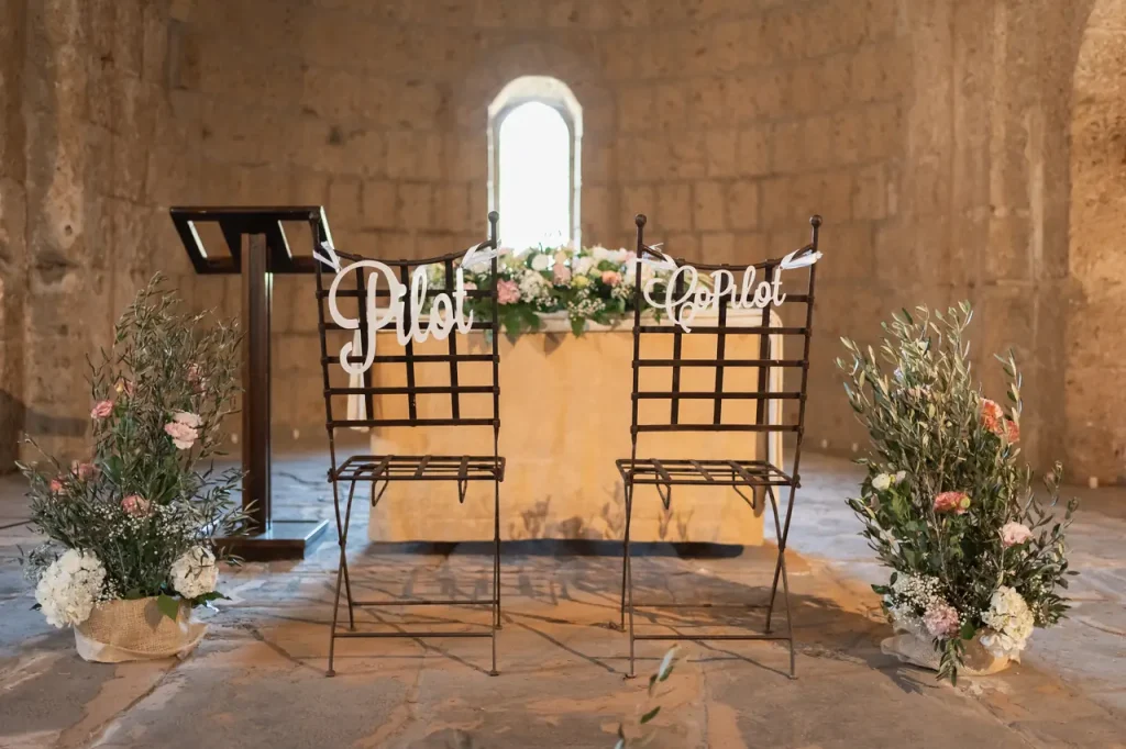 Due sedie di metallo con le scritte "pilota" e "copilota" sono di fronte a un altare decorato con fiori in una cappella con pareti in pietra, catturando perfettamente l'essenza dell'allestimento cerimonia.