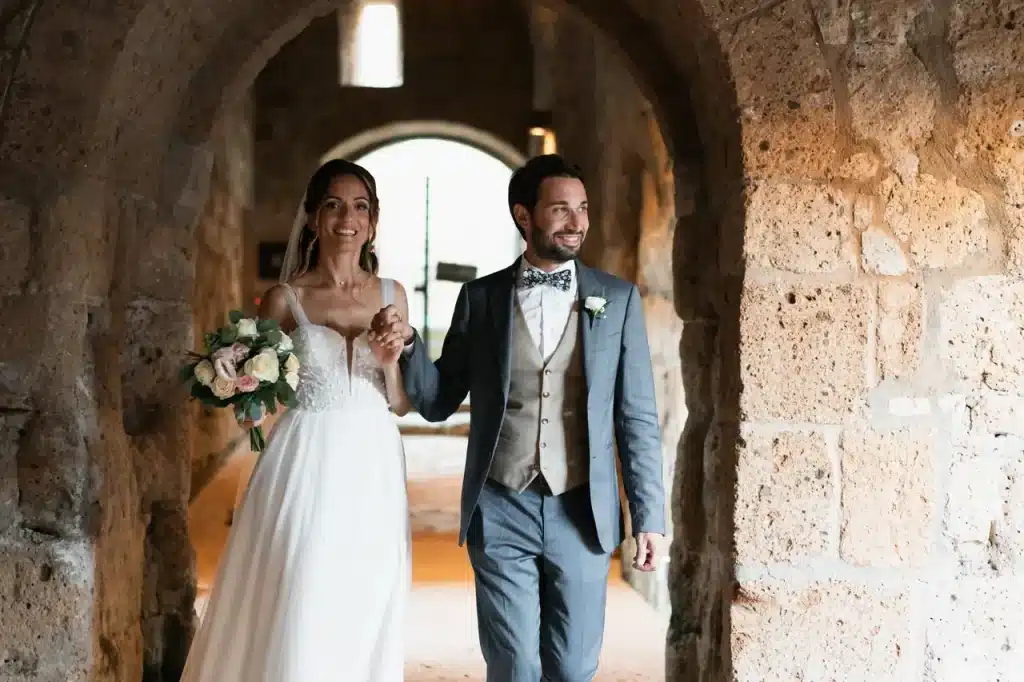 Una sposa e uno sposo camminano mano nella mano attraverso un arco di pietra. La sposa tiene in mano un bouquet di fiori.