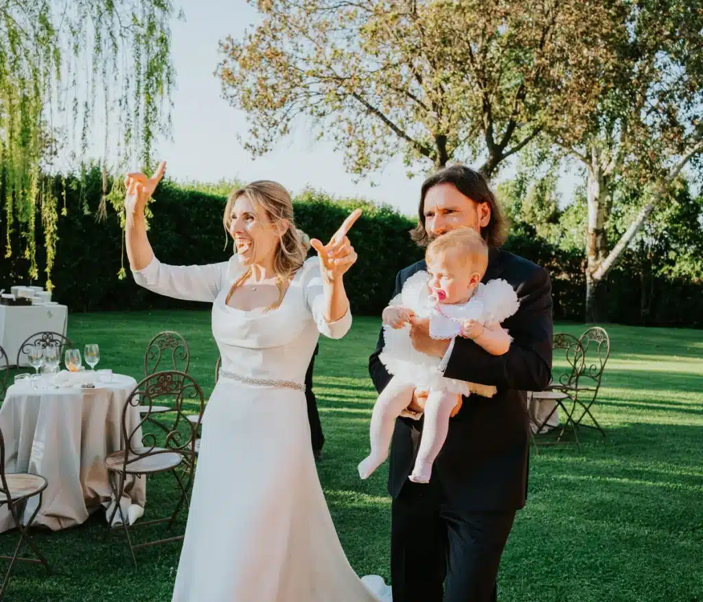 A woman in a wedding dress is gesturing enthusiastically whilst a man in a suit and tie is holding a child in his arms; they are outdoors on a lawn, with tables and trees in the background.