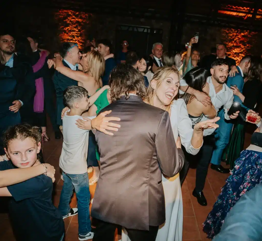 A group of people are dancing in close contact at an indoor event, with subdued lighting and several couples embracing on the dance floor.