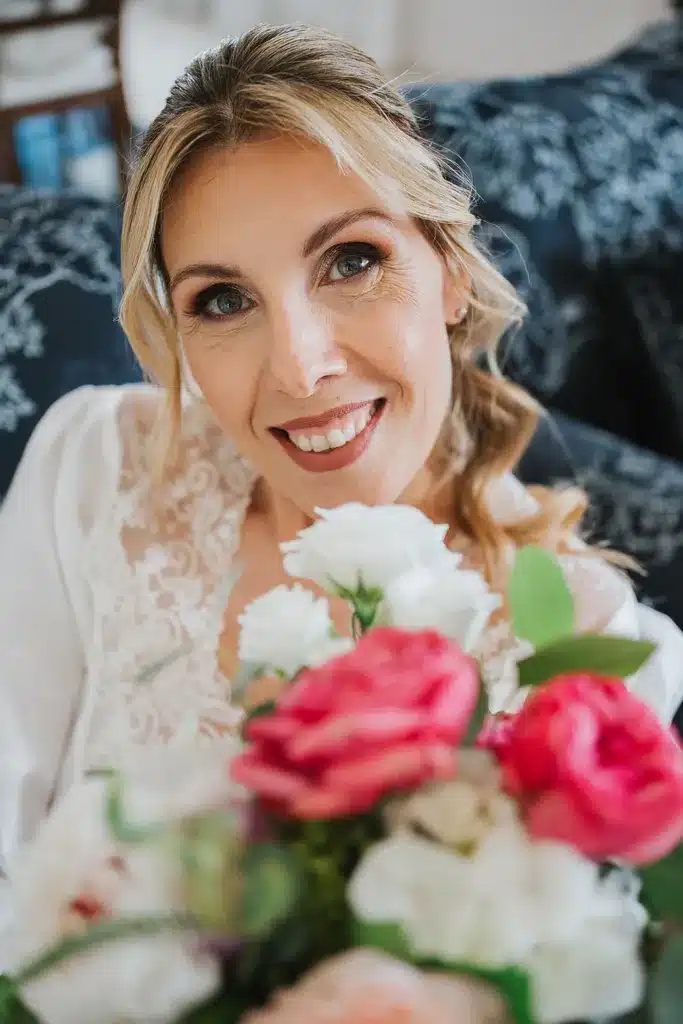 A blonde woman smiles at the camera, holding a bouquet of pink and white flowers and wearing a white lace top.
