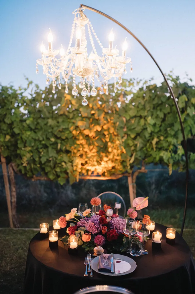 Tavolo da pranzo all’aperto con centrotavola floreale, candele e stoviglie, sistemato sotto un lampadario di cristallo con tralci di vite sullo sfondo al crepuscolo.